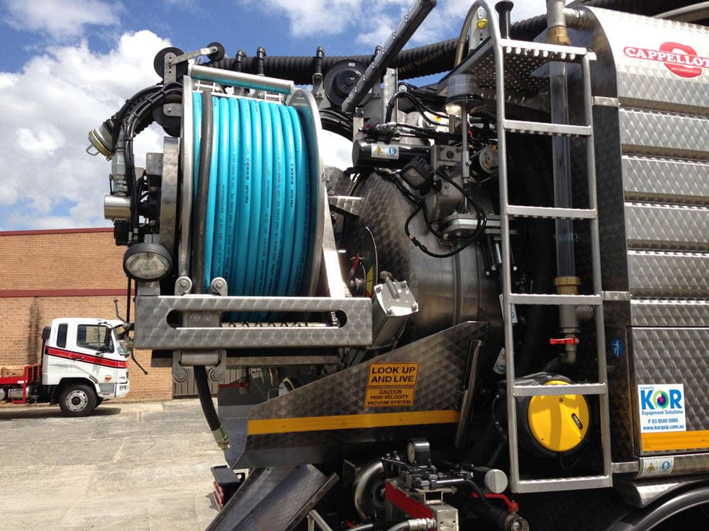 Close-up of a large industrial vacuum truck with a mounted blue hose reel, metal tank, ladder, and various control mechanisms. A white truck is parked in the background.