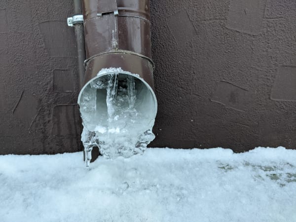 A brown metal drainpipe with icicles and ice buildup at the opening, attached to a textured wall above snow-covered ground—a clear reminder of the importance of preventing frozen pipes in winter.