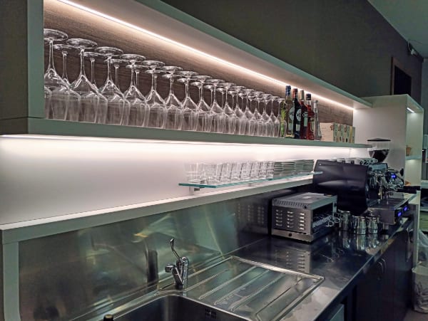 A modern bar setup with rows of glasses, bottles of liquor, a coffee machine, and various bar equipment on stainless steel shelves and counter, complete with accessible plugs for all your devices.