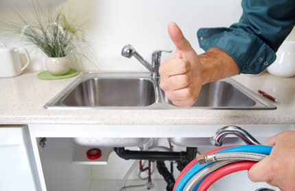 Person gives a thumbs up in front of a kitchen sink while holding plumbing hoses, indicating successful work fixing a slow drain.