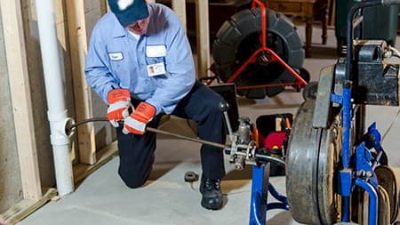 A plumber uses a drain snake machine to clear a pipe in a basement. We're hiring—join our team and start building your plumbing career today!.