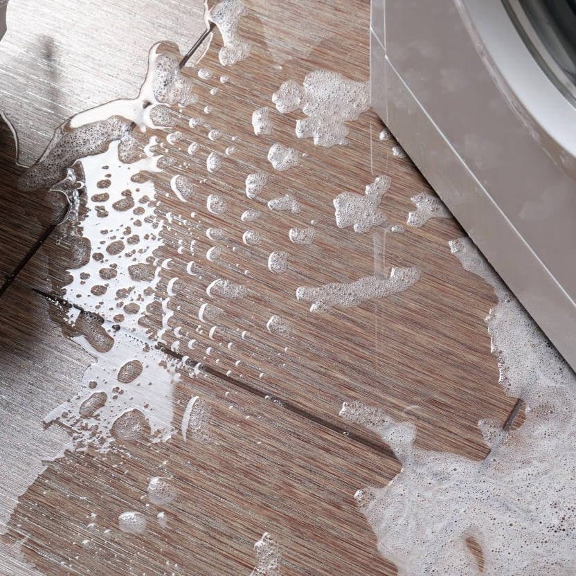 Soapy water spilling onto a wooden floor from the side of a washing machine, creating bubbles and puddles—a clear sign that drain cleaning may be needed.