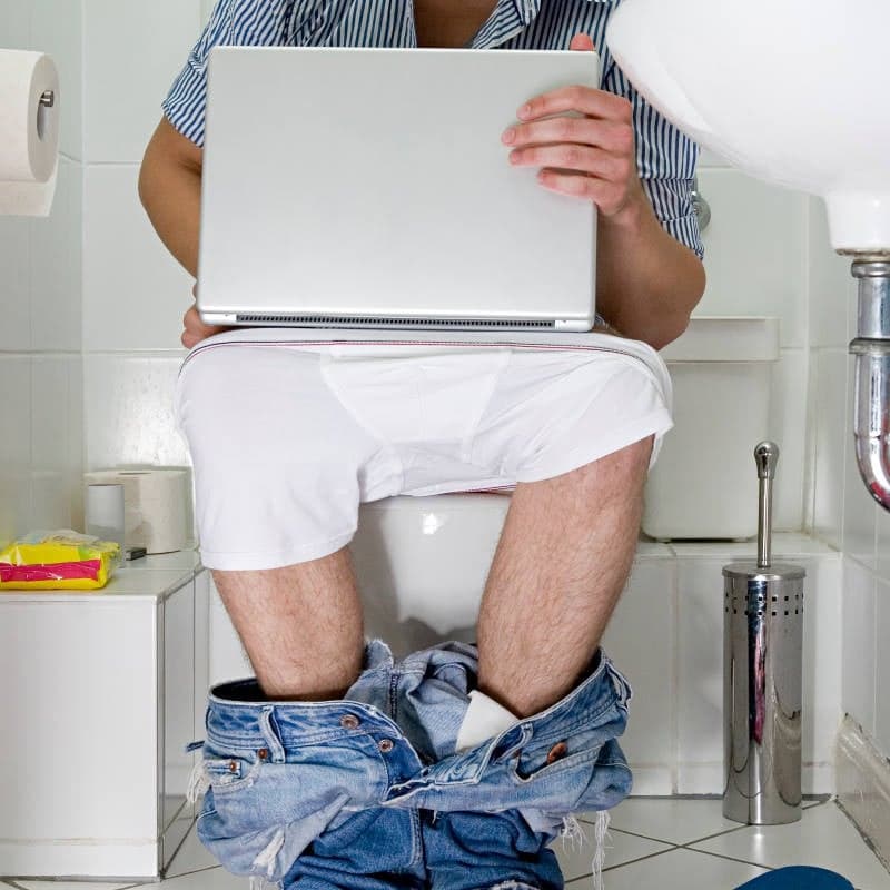 A person sits on a toilet with pants and underwear down, using a laptop in the bathroom—perhaps searching plumbing services to fix clogged toilets or handle urgent toilet repair needs.