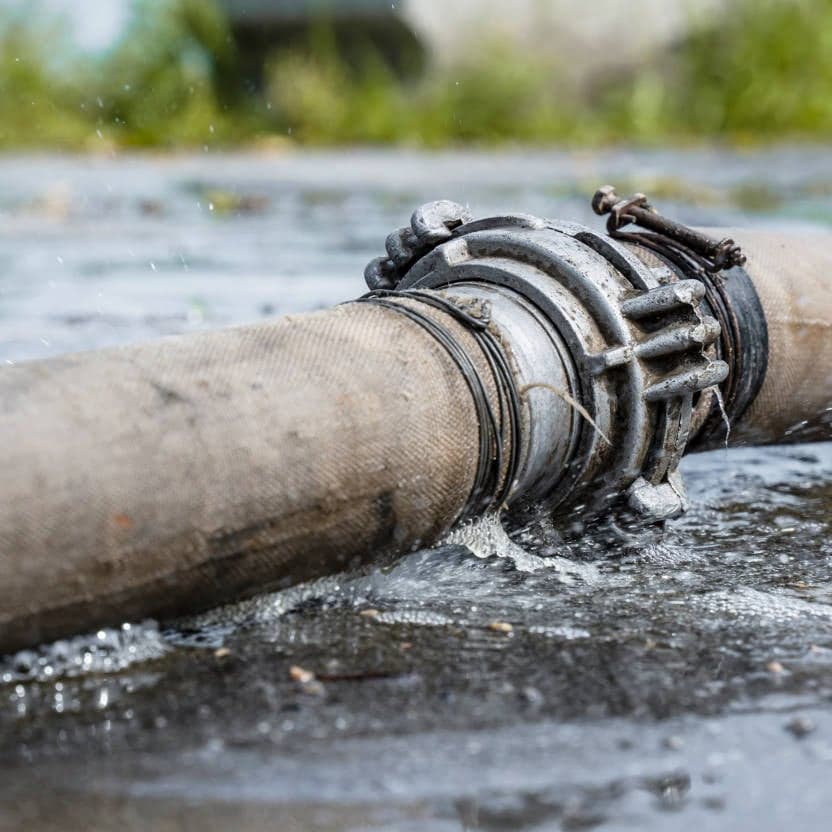 A large industrial hose is jetting water from a leak at the metal connector, causing water to pool on the ground.