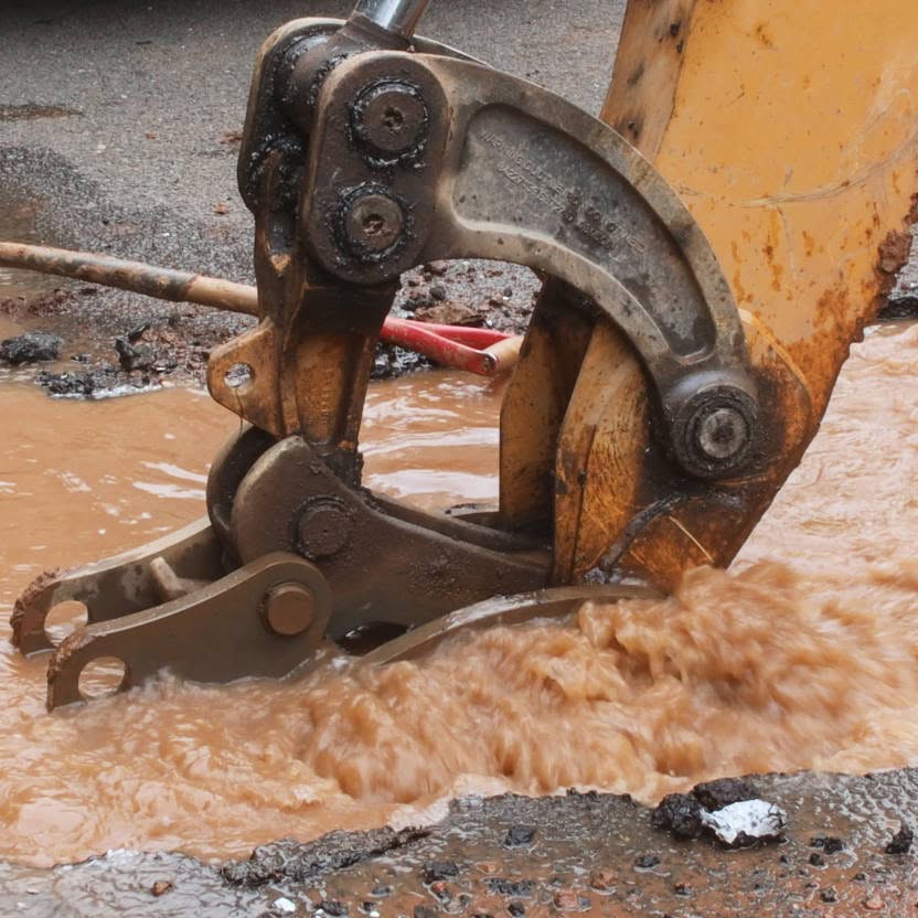 A close-up of an excavator bucket digging into muddy water at a construction site, possibly verifying sewer lines before repairs.