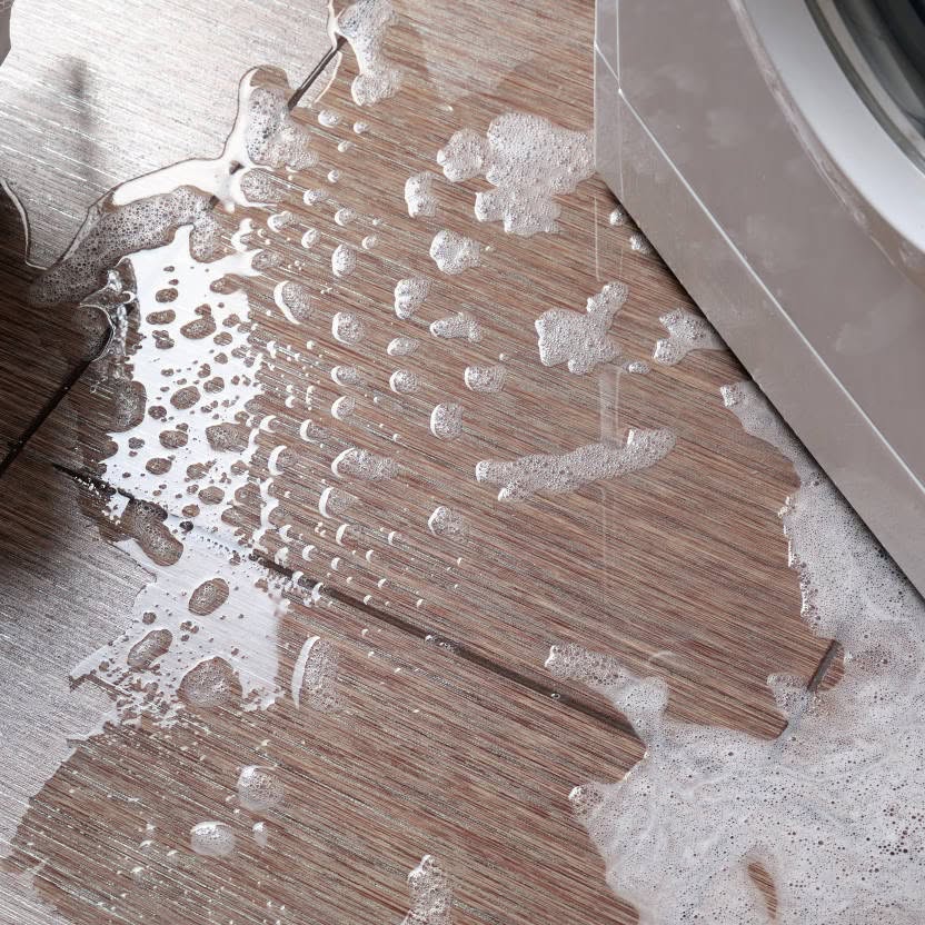 Soapy water spilling onto a wooden floor from the side of a washing machine, creating bubbles and puddles—a clear sign that drain cleaning may be needed.